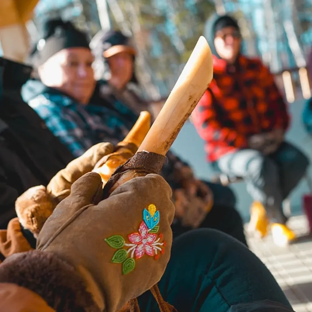 Close-up of a person outdoors wearing beaded mittens while holding a carved bone tool, with others seated in the background