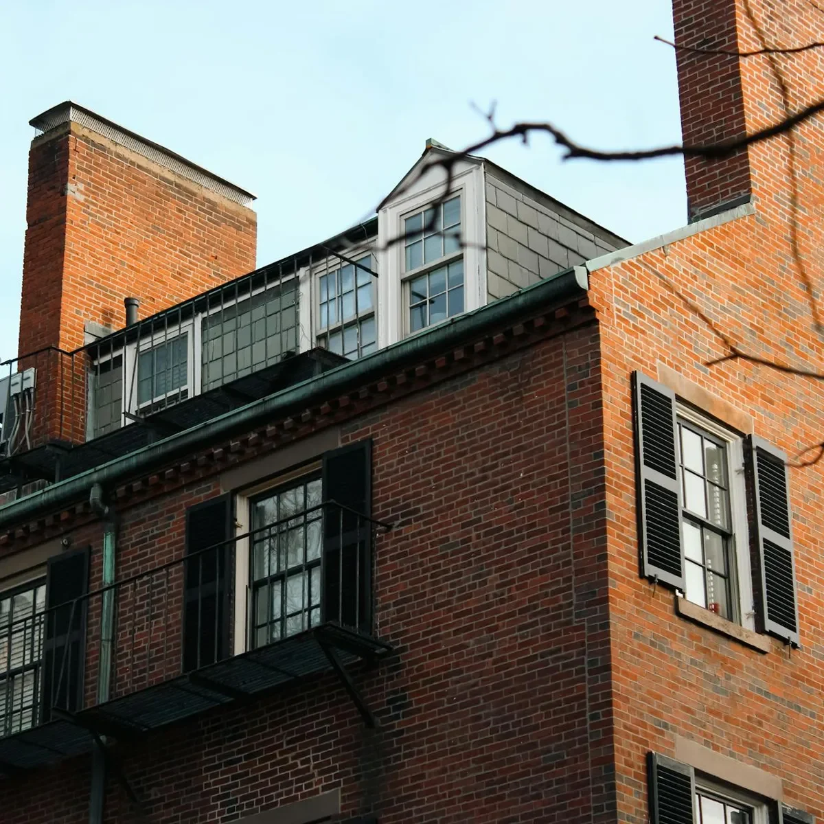 Brick residential building with black shutters, rooftop windows, and a metal fire escape under a clear sky