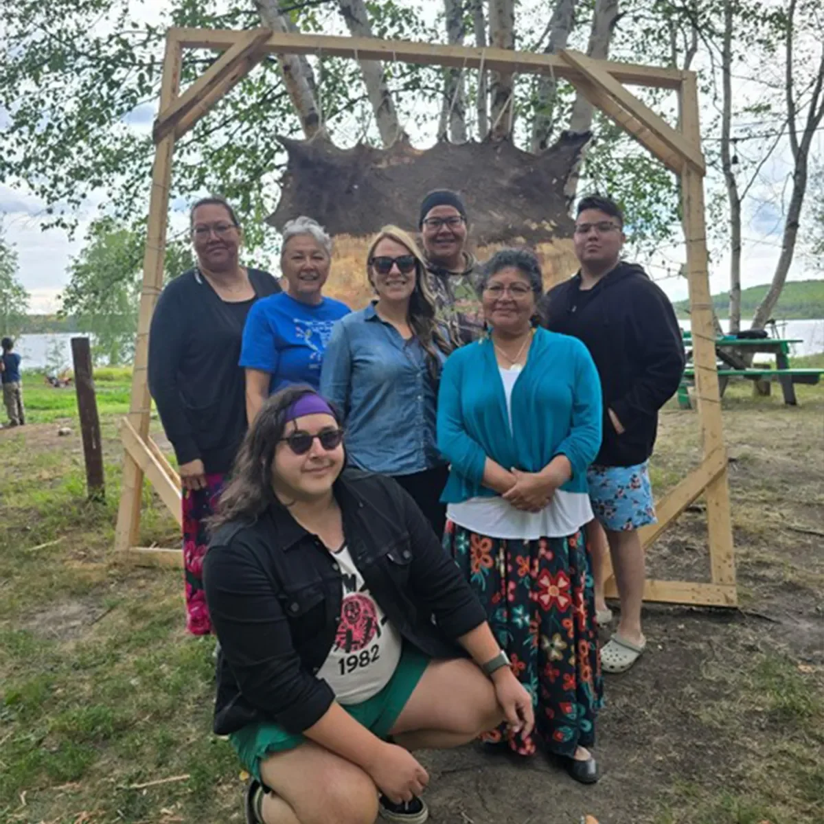 Group of people gathered outdoors under a wooden structure with a stretched animal hide, smiling and posing together.
