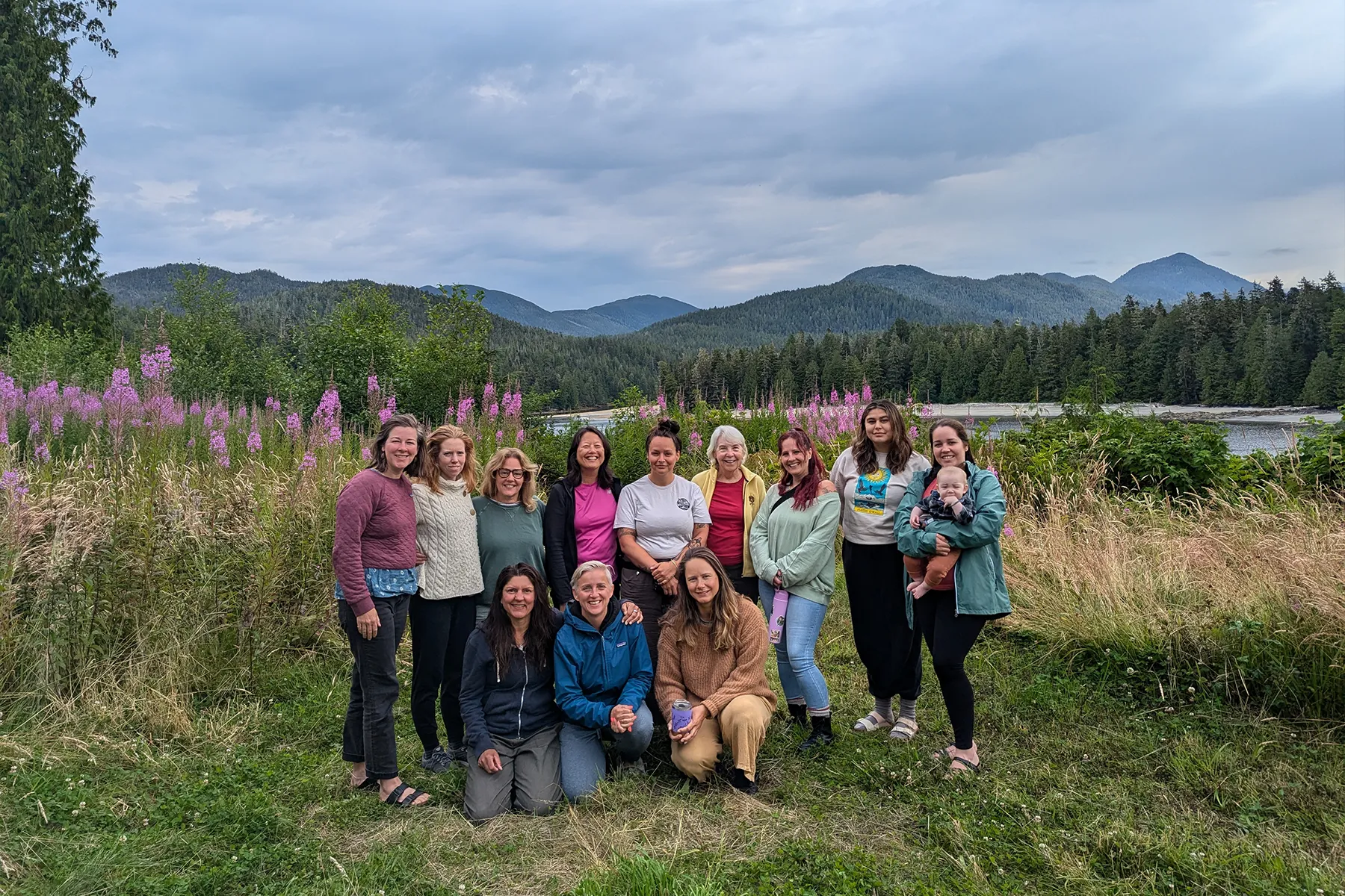 Group of people posing outdoors in a grassy field with mountains and trees in the background.